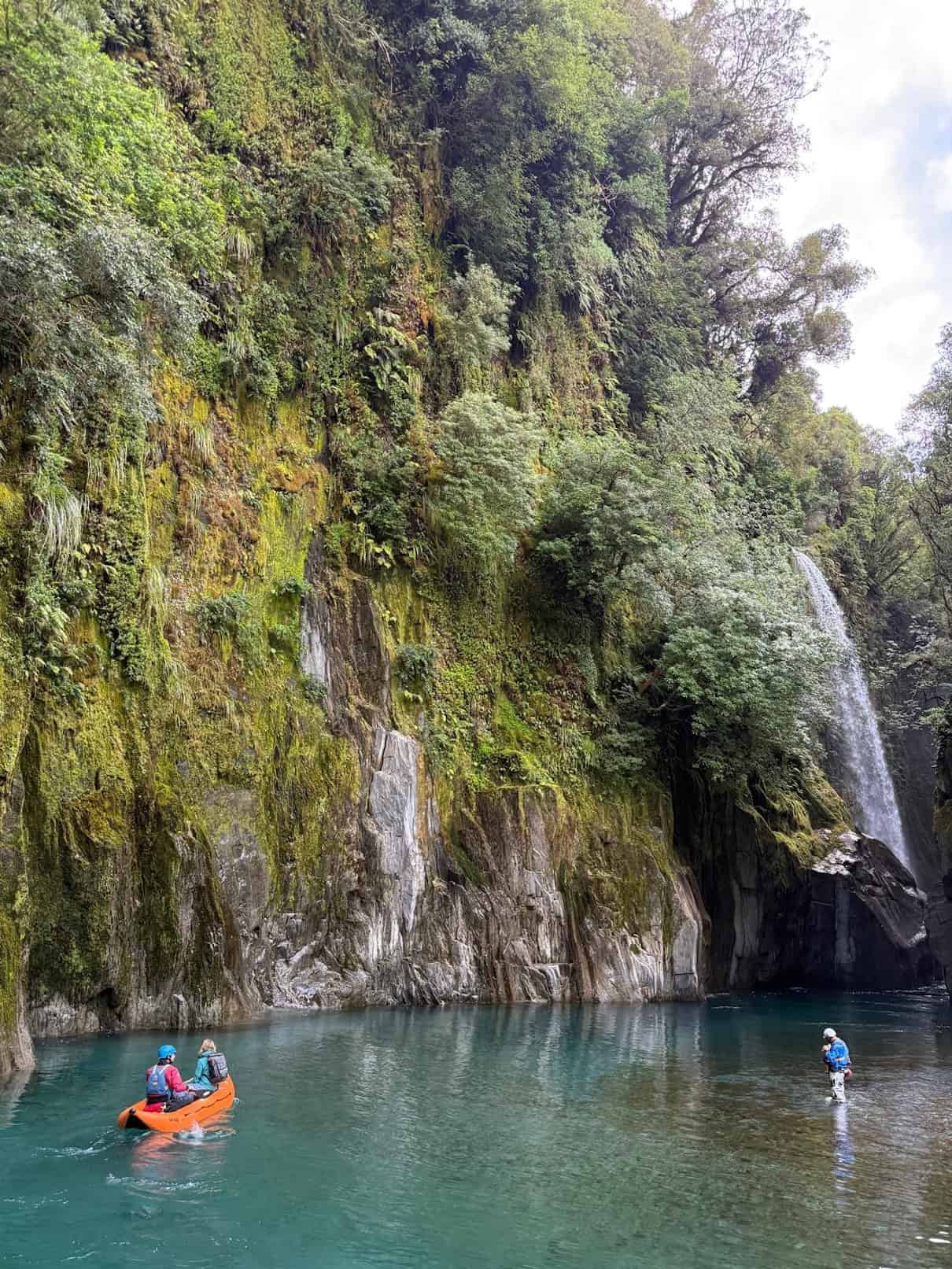 Two people in orange inflatable kayak next to moss-covered rock wall