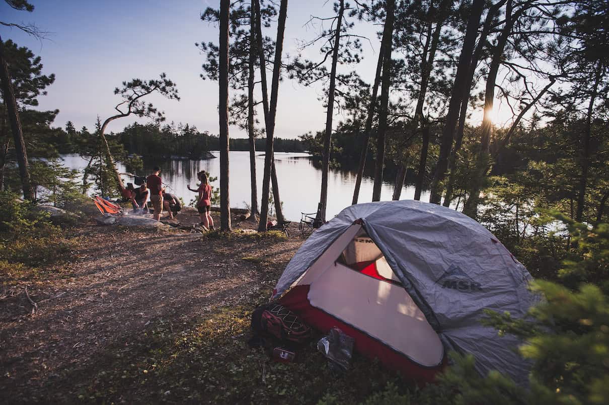 People gathered on rocky campsite among trees with tent set up in foreground.