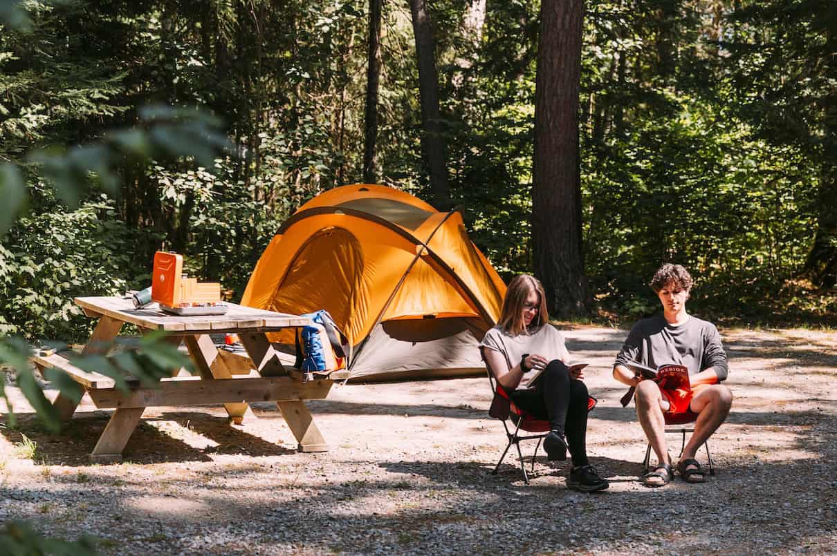 Two people sitting on camp chairs with tent and picnic table behind them.