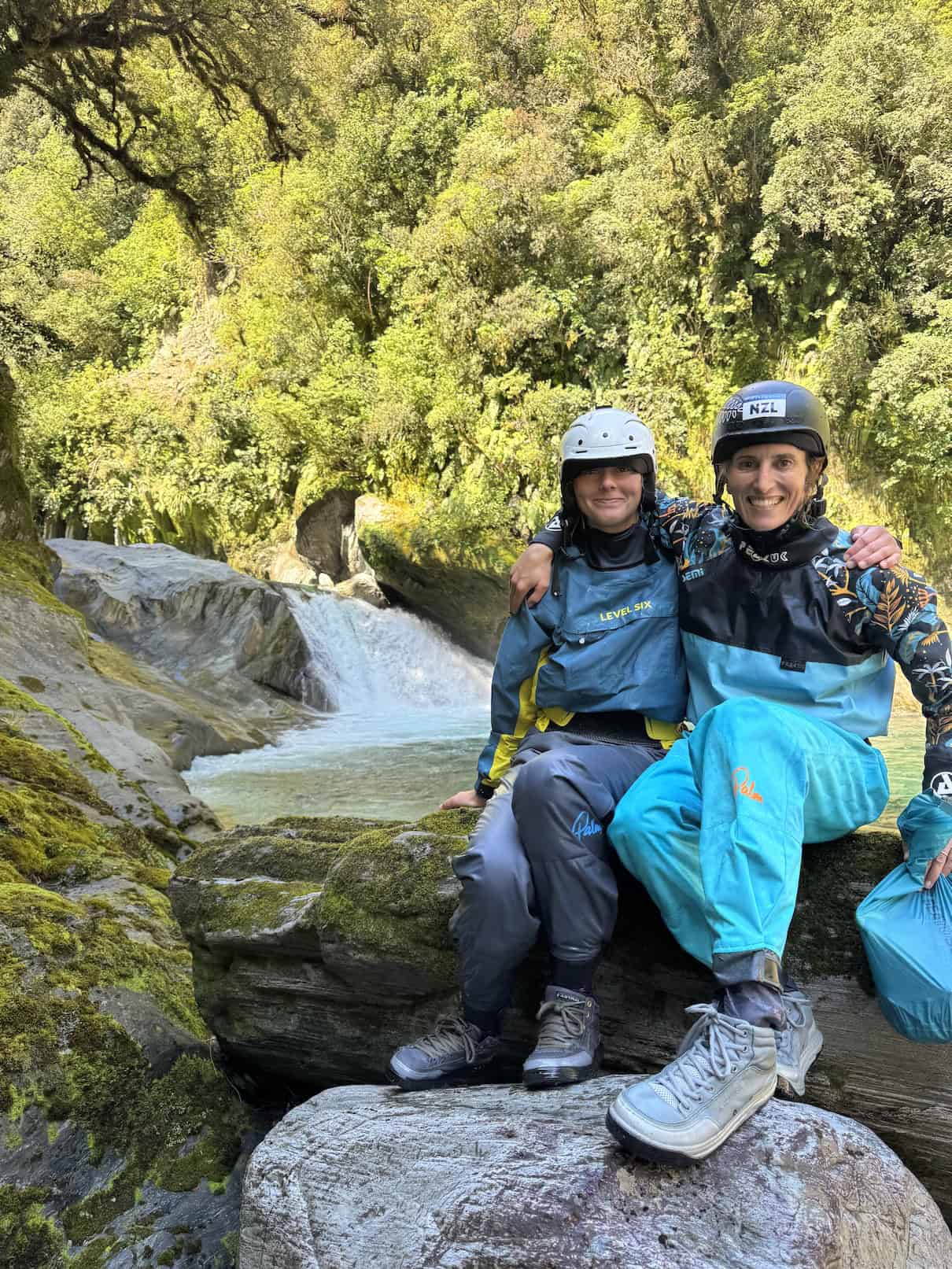 Two women in drysuits sitting on rock in front of rapid
