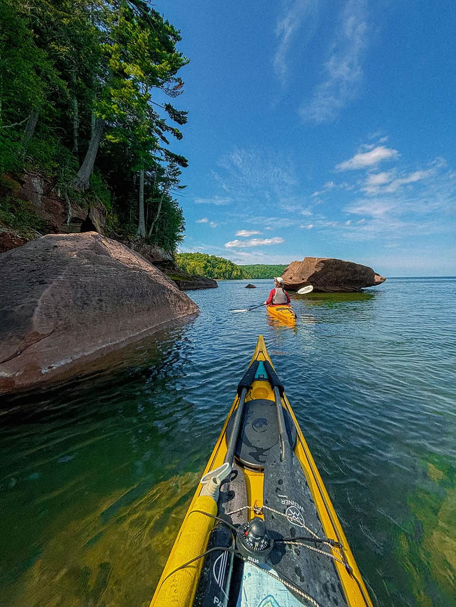 bow shot as two women paddle along a rocky, verdant shoreline