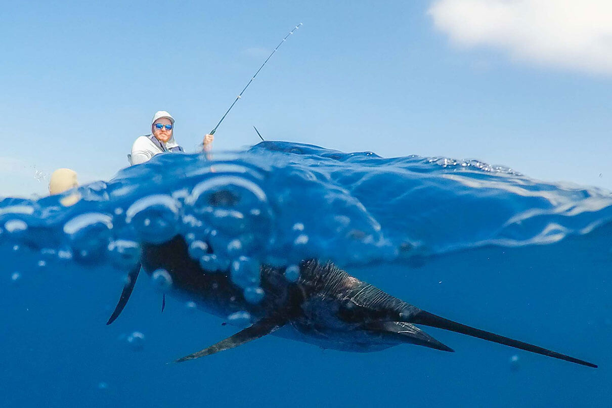 man hooks a sailfish under the blue water while fishing from a kayak under blue skies