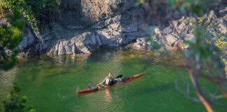 What’s In Editor Maddy Marquardt’s Sea Kayak Day Trip Guiding Kit Paddling Magazine Editor Maddy Marquardt in her sea kayak on a trip with vivid green water, rocks and surrounding foliage in the foreground