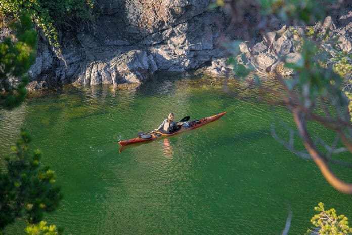 Paddling Magazine Editor Maddy Marquardt in her sea kayak on a trip with vivid green water, rocks and surrounding foliage in the foreground