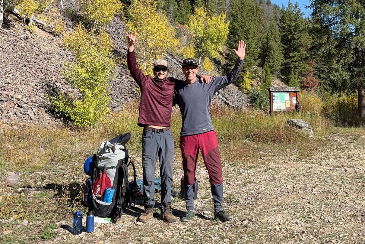 Kayakers Micah Barker and Jeff Landers stand and wave with outdoor gear at a trailhead