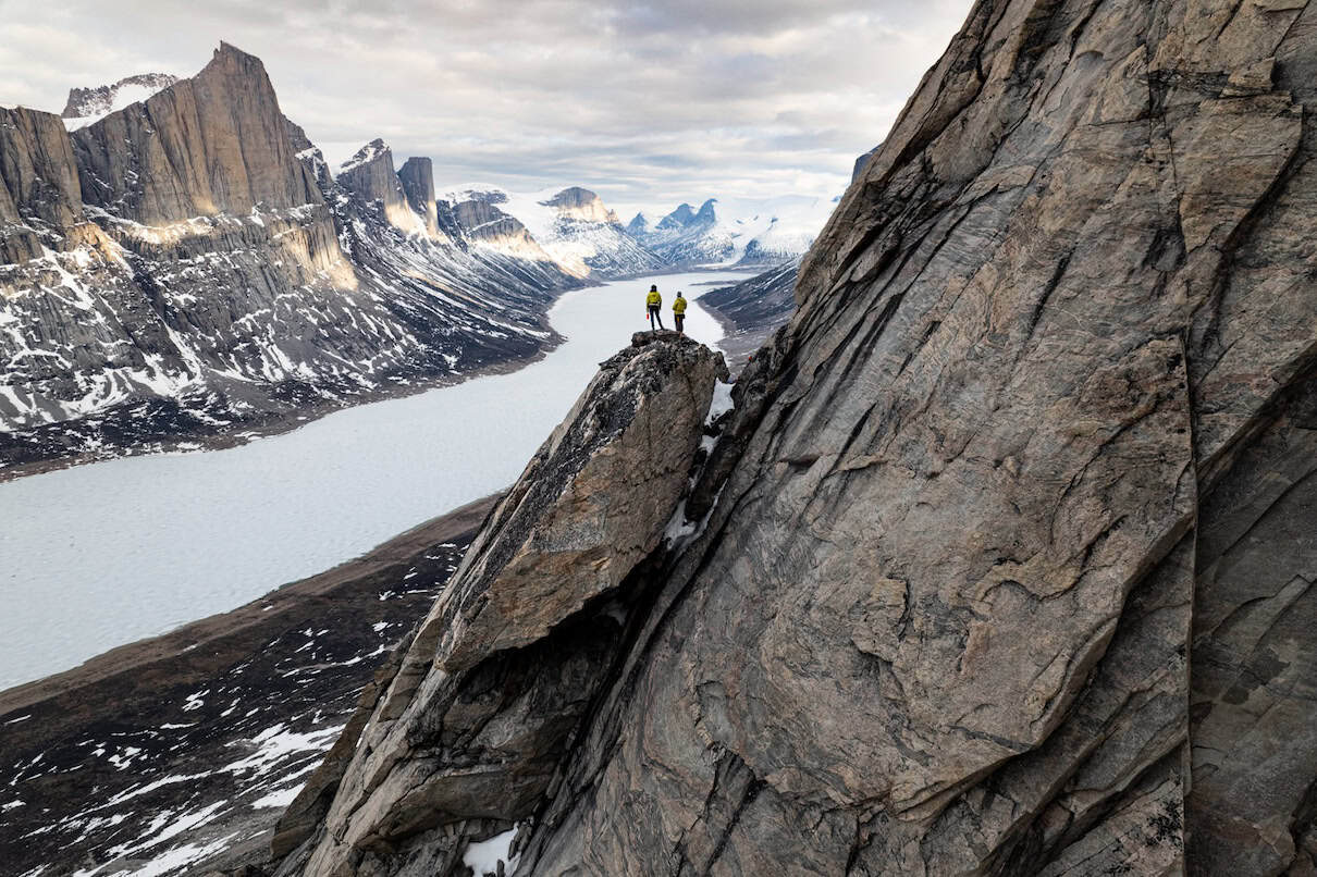 Two people standing atop rocky spire on side of mountain looking down at frozen ocean below