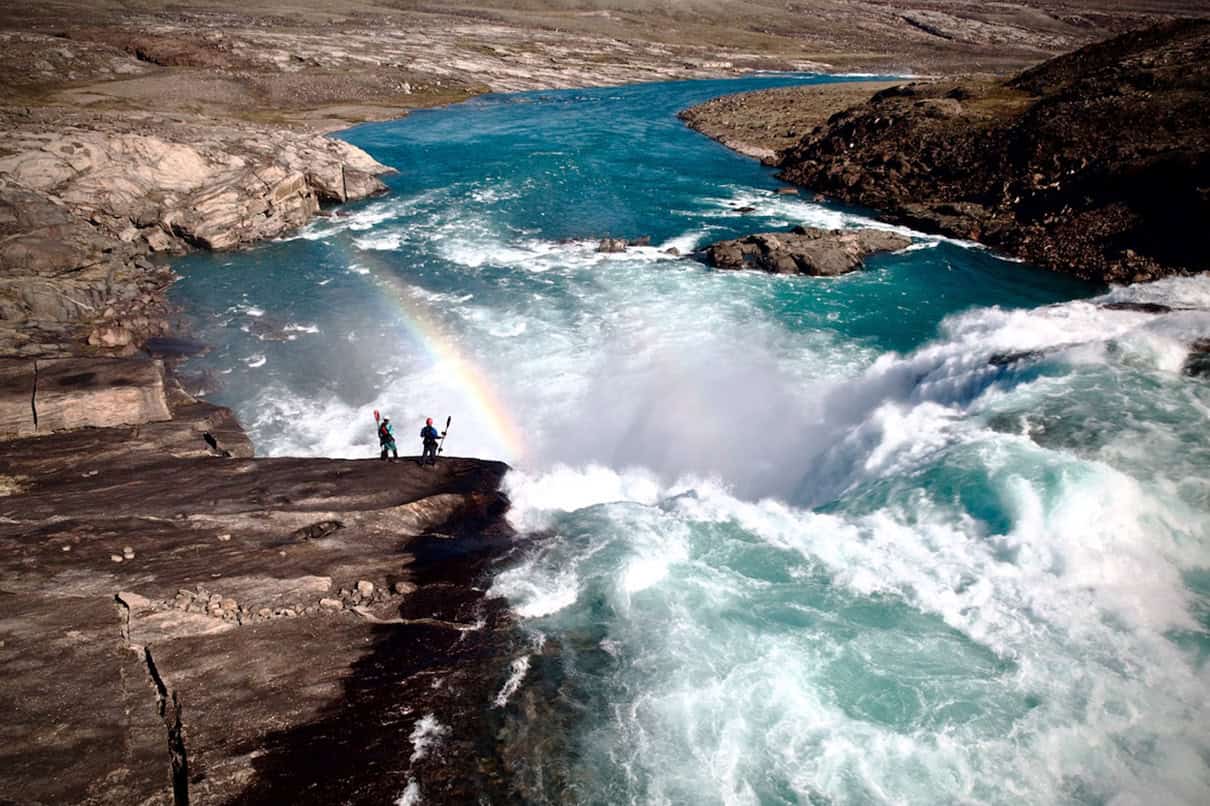 Two people holding kayak paddles standing on rocky ledge overlooking waterfall with rainbow