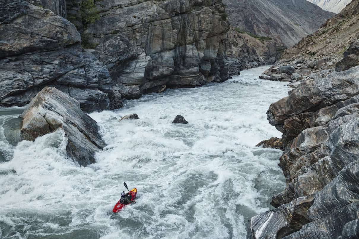 Person in red whitewater kayak going down big rapid with massive boulders and rocky banks