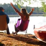 Best Camp Chairs For 2026 Two people sitting in camp chairs near a lake with canoe in foreground.