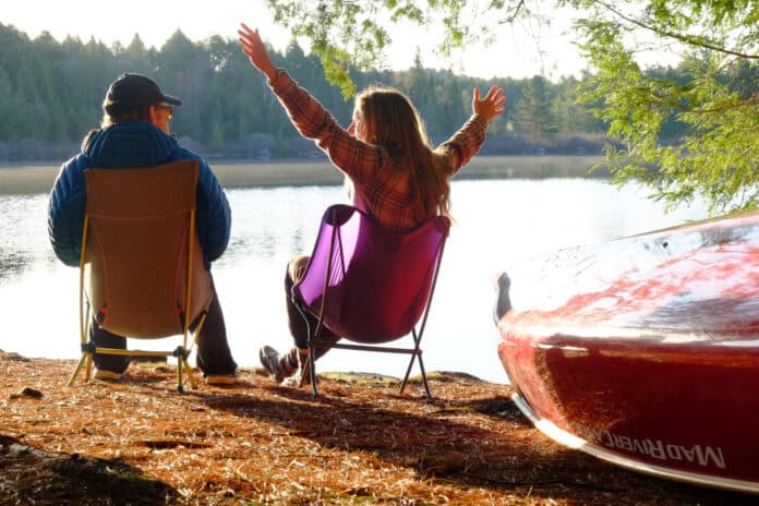 Two people sitting in camp chairs near a lake with canoe in foreground.