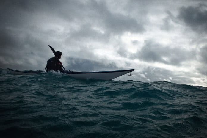 person sea kayaks on swell under dark, cloudy skies