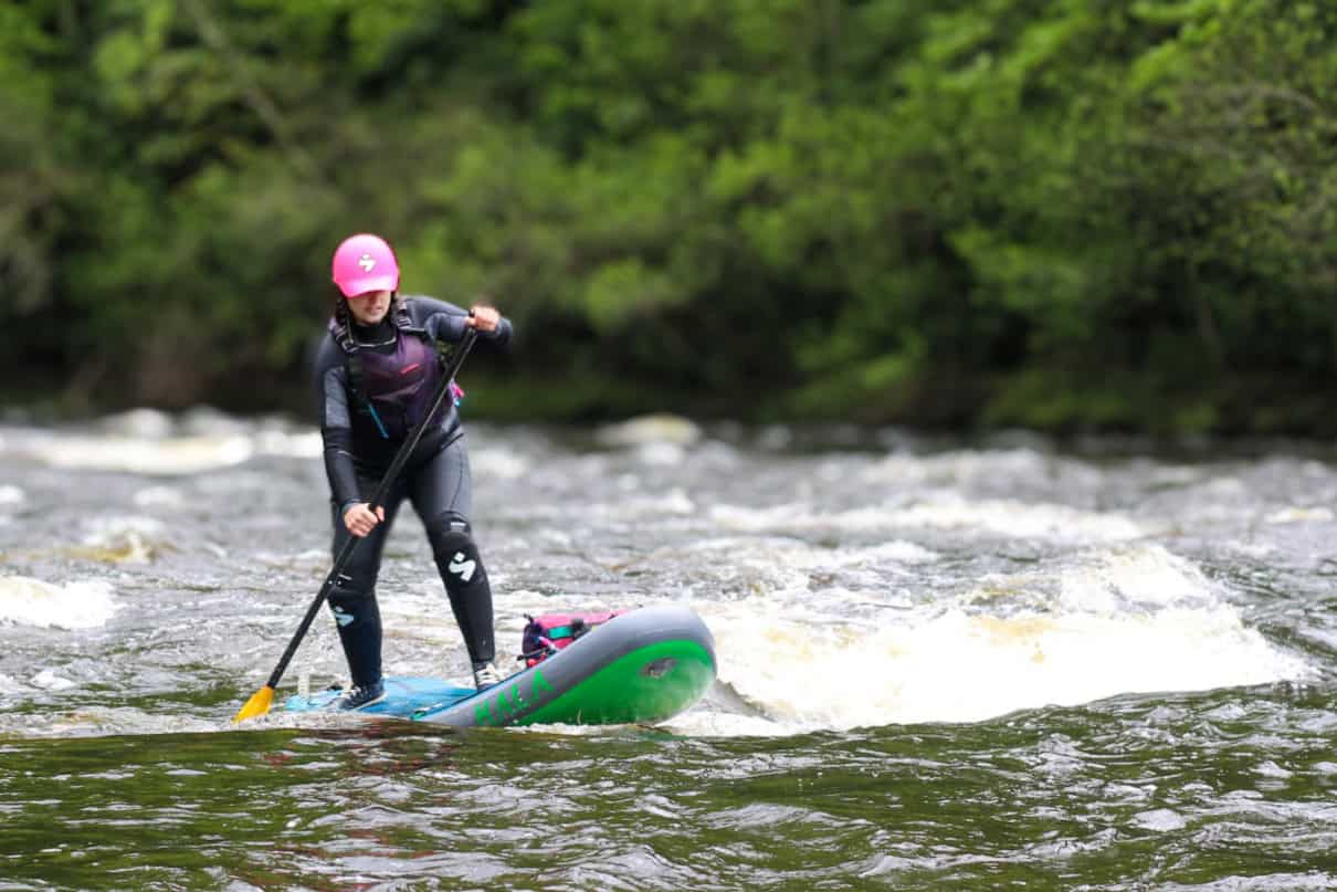 Standup paddleboarder surfing a river wave while wearing a wetsuit.